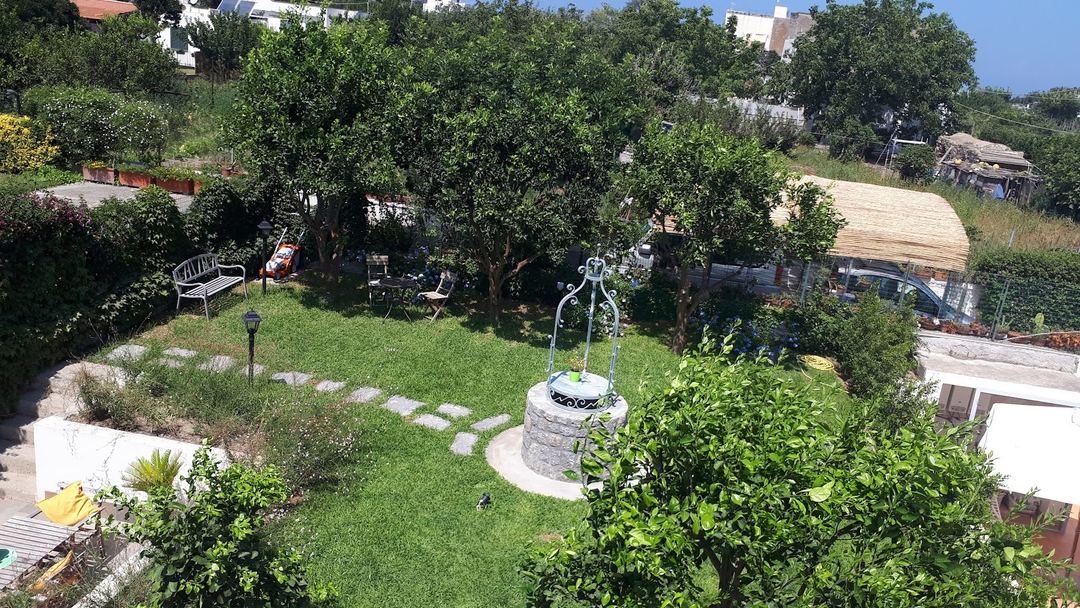 Aerial view of a Mediterranean-style garden with mature trees, seating areas, a stone well feature, and manicured lawn surrounded by residential buildings.