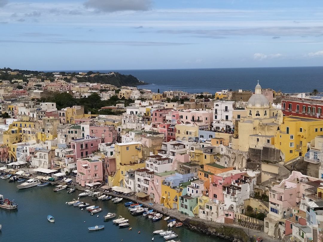 Panoramic view of a colorful Mediterranean coastal town with pastel-colored buildings, a domed church, and boats in a turquoise harbor.