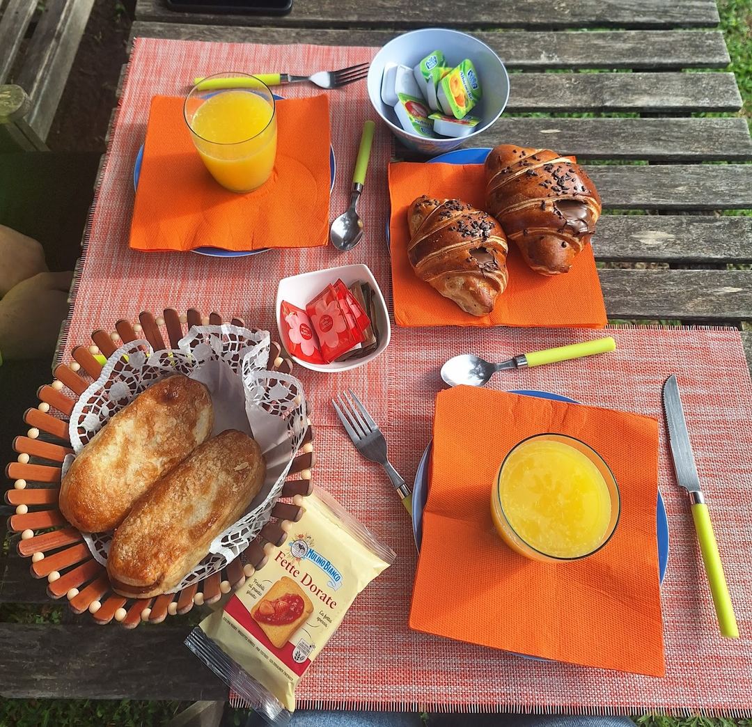 Outdoor breakfast setting featuring fresh pastries, orange juice, cheese, tomatoes, and bread rolls arranged on an orange napkin-lined table.