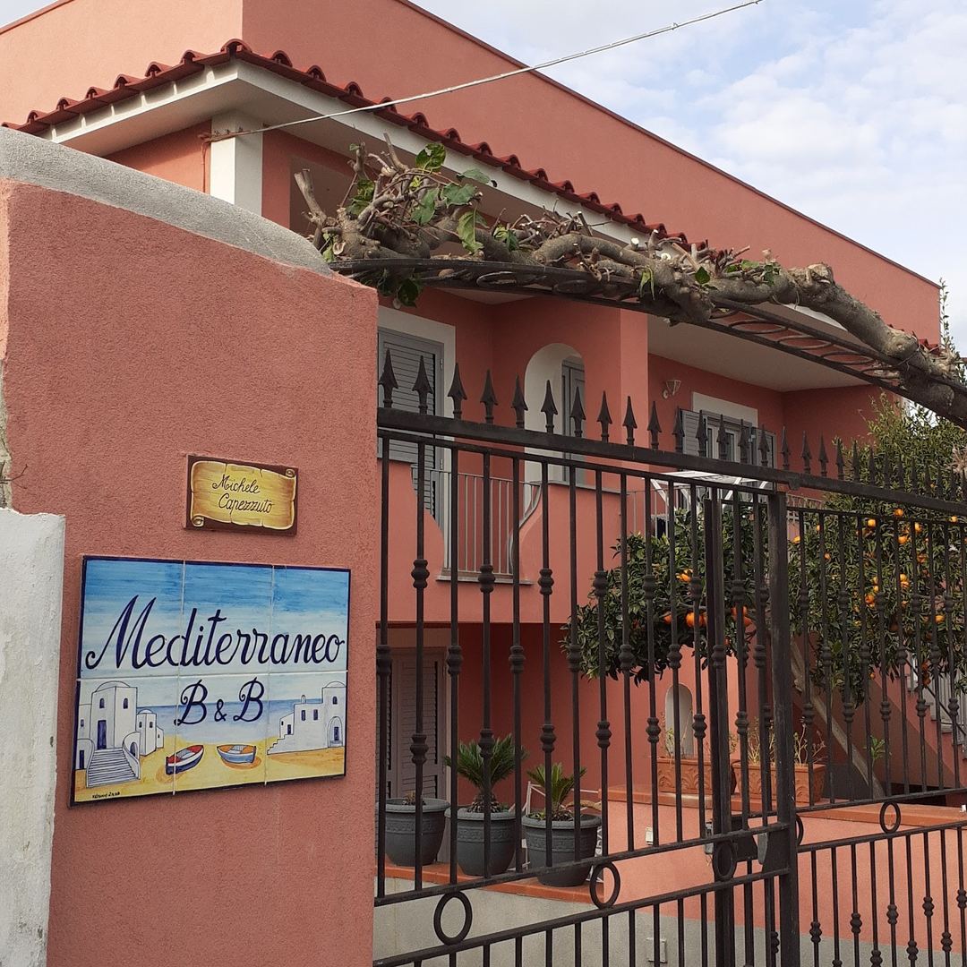 The terracotta-colored facade of B&B Mediterraneo with decorative signage, wrought-iron gate, and rustic pergola overhead.