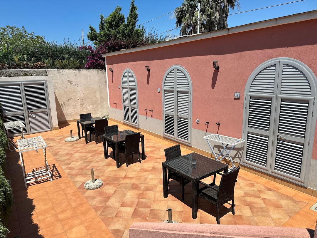 A sunny terracotta-tiled courtyard with black wicker furniture and arched louvered doors on a coral-pink building at B&B Mediterraneo.