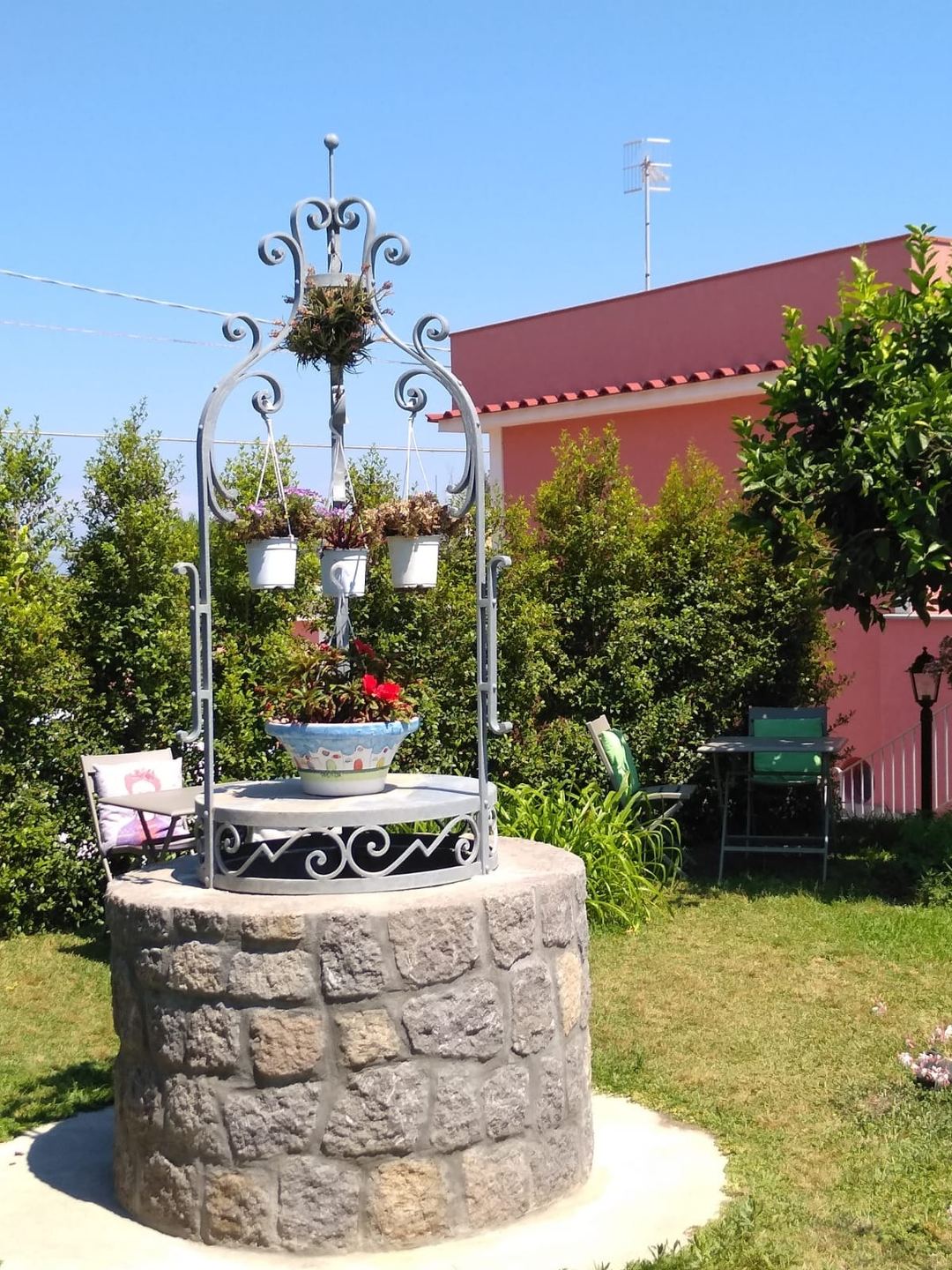 A decorative ornamental well structure with wrought iron scrollwork and hanging flower baskets in the garden of a Mediterranean-style property.