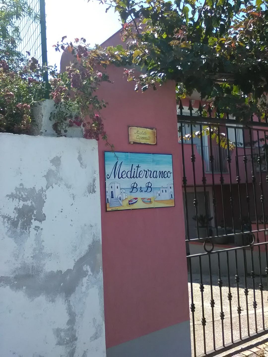 The terracotta-colored exterior entrance of B&B Mediterraneo with a decorative sign and iron gate, framed by climbing vines.